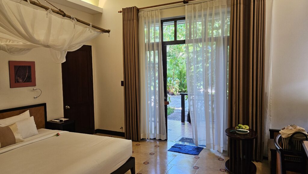 Spacious guest room at Bamboo Village Resort with a canopy bed, garden view through large glass doors, and tropical ambiance.