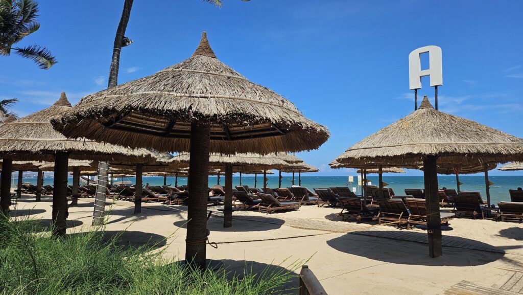 hatched beach umbrellas and wooden sun loungers lined up on the sandy beach at Bamboo Village Resort, facing the ocean.