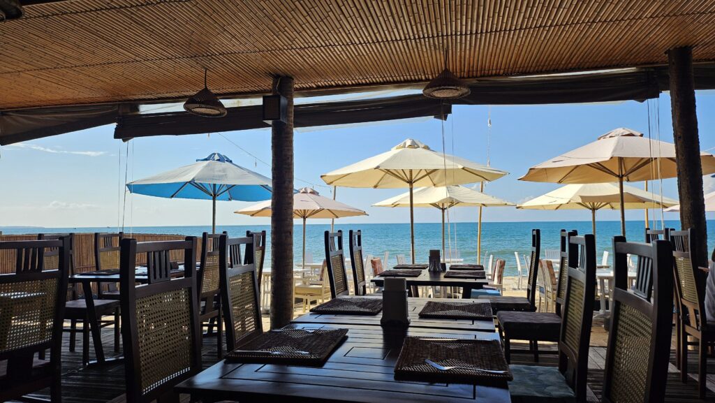 Beachfront restaurant at Bamboo Village Resort with wooden chairs and tables overlooking the ocean under parasols.
