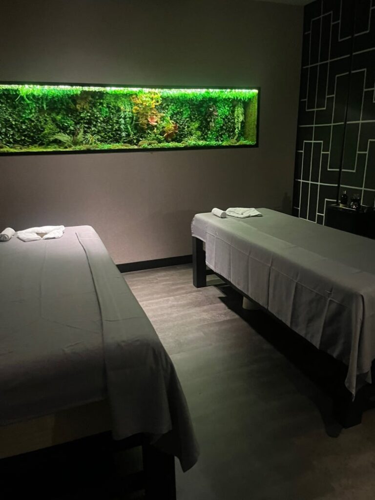 A modern, dimly-lit massage room with two massage beds and a lush green aquarium-style wall feature.