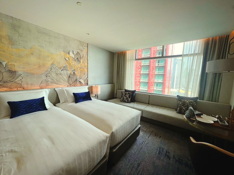 A spacious twin bedroom in Chatrium Grand Bangkok hotel with large windows and a view of high-rise buildings.
