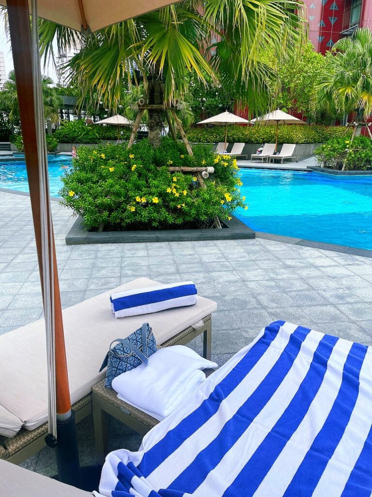 A striped towel and sunbed by the poolside at Chatrium Grand Bangkok with lush greenery and palm trees.