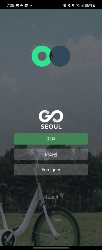 Main login screen of the Seoul public bike service app "Ttareungi." At the top is a loading animation with two overlapping circles. Below, there are three login options: "Member," "Non-member," and "Foreigner." The background image shows a white public bicycle on a grassy field.