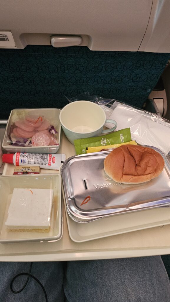 Vietnam Airlines economy class in-flight meal tray including bread roll, salad, dessert, instant rice topping sauce, and tea packet before opening the main course.
